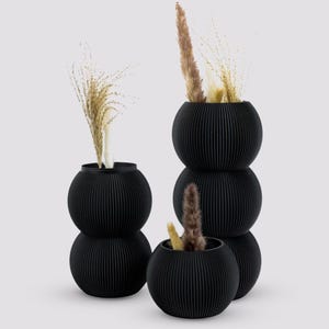 May include: Three black, round, ribbed planters with dried flowers. The planters are stacked on top of each other.