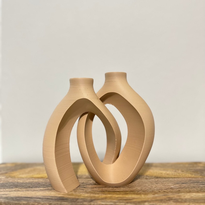 May include: Two beige ceramic vases with a unique, abstract design. The vases are connected and have a curved, open shape.