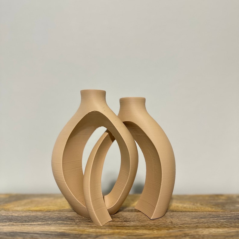 May include: A set of two beige ceramic vases with an abstract, modern design. The vases are connected and have a unique, open shape.