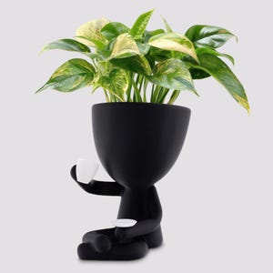May include: A black planter in the shape of a person, holding a small white teacup and saucer. The planter is filled with a green and yellow variegated plant. The design is modern and whimsical, suitable for indoor decor.