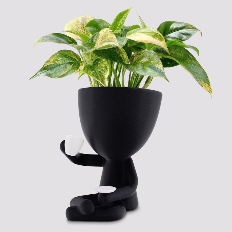 May include: A black planter in the shape of a person, holding a small white teacup and saucer. The planter is filled with a green and yellow variegated plant. The design is modern and whimsical, suitable for indoor decor.