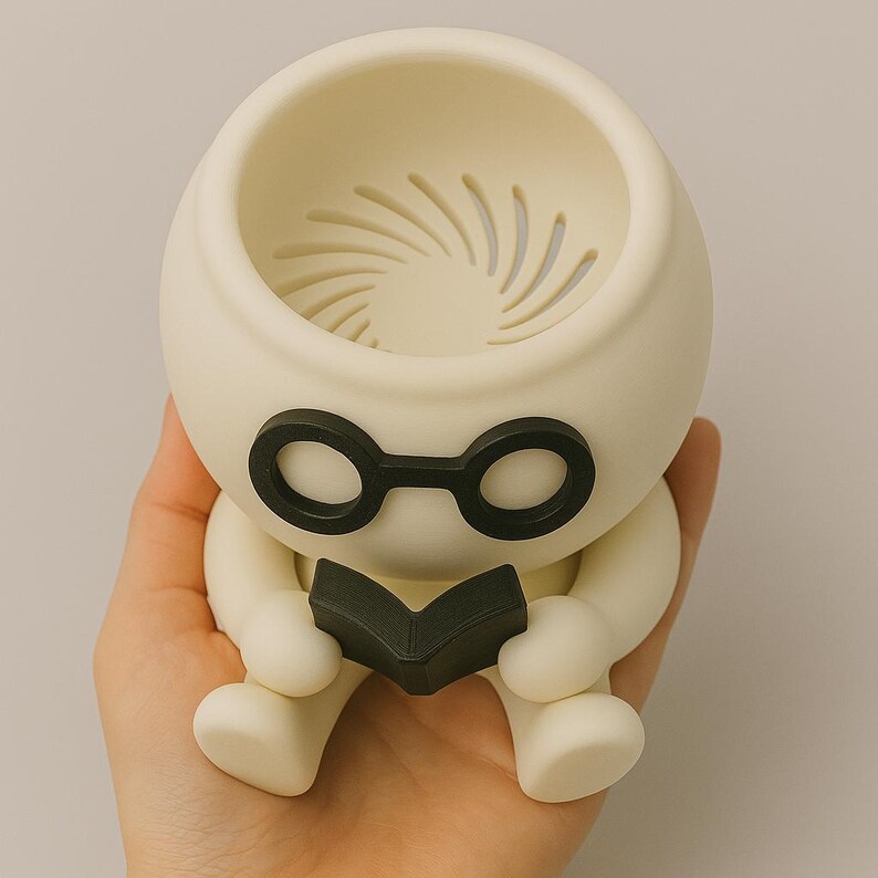 May include: A white ceramic planter shaped like a person wearing glasses and holding a book. The planter has a drainage hole in the top.