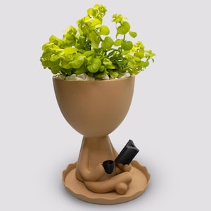 May include: A brown ceramic planter shaped like a person sitting in a meditative pose. The planter has a wide, rounded top and a small, round base. The person is holding a small, black book in one hand and a small, black cup in the other. The planter is filled with green plants.