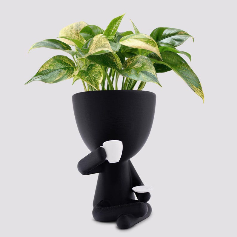 May include: A black ceramic planter shaped like a person, holding a white teacup and saucer. The planter is filled with a green and yellow variegated plant. The design is whimsical and modern, suitable for indoor decor.