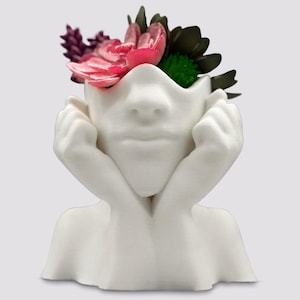 May include: White ceramic planter shaped like a person's head with hands holding their chin. The planter has a flower pot in the head area with pink and green flowers.