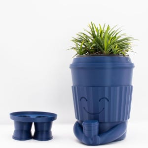 May include: A blue ceramic planter shaped like a coffee cup with a smiling face. The planter has a ribbed design and is holding a smaller coffee cup. A green plant is growing in the planter.