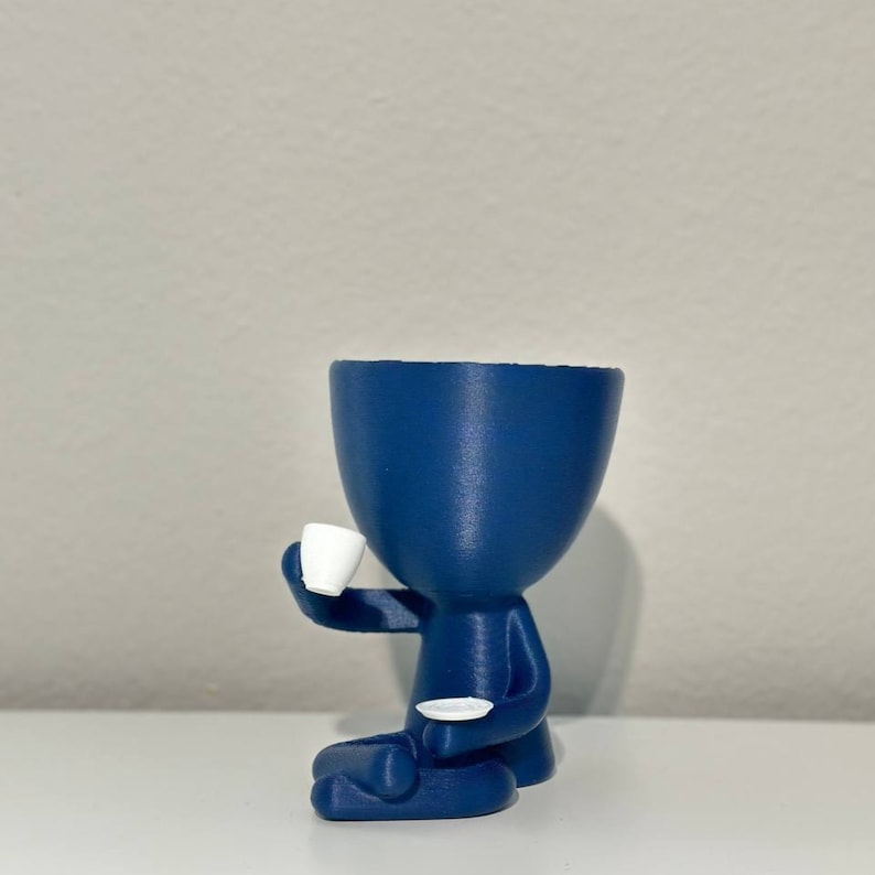 May include: A navy blue planter shaped like a person, holding a small white teacup and saucer. The planter has a large opening at the top for plants. The figure is in a seated position, with its legs crossed.