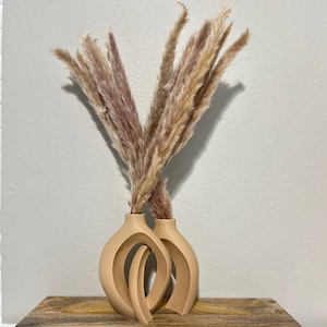 May include: Two beige ceramic vases with an abstract design, holding a bouquet of dried pampas grass.