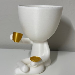 May include: A white, 3D-printed planter shaped like a person, holding a gold-colored teacup and saucer. The figure is seated with its legs crossed. The planter has a large opening for plants.