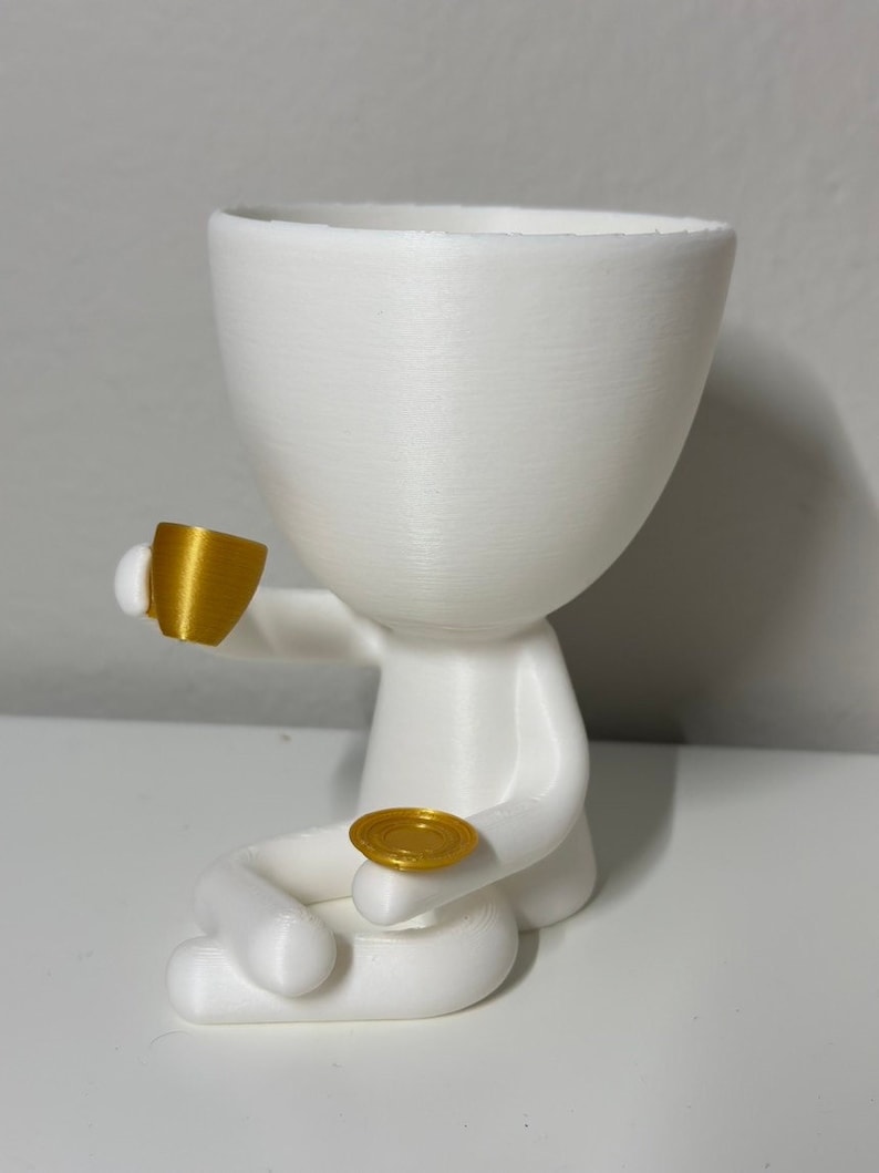 May include: A white, 3D-printed planter shaped like a person, holding a gold-colored teacup and saucer. The figure is seated with its legs crossed. The planter has a large opening for plants.