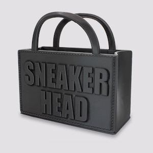 May include: Black rectangular handbag with two handles. The words "SNEAKER HEAD" are embossed on the front. The bag is made of a matte material and has a stitched border.