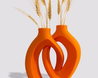 Könnte beinhalten: Zwei orangefarbene Keramikvasen mit modernem, abstraktem Design. Die Vasen sind ineinander verschlungen und haben eine geschwungene, offene Form. Die Vasen sind mit getrockneten Weizenhalmen gefüllt.