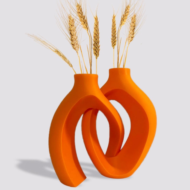 May include: Two orange ceramic vases with a modern, abstract design. The vases are intertwined and feature a curved, open shape. The vases are filled with dried wheat stalks.