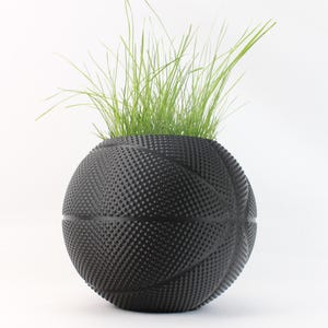 May include: A black basketball-shaped planter with green grass sprouting from the top. The planter has a textured surface with the typical lines and dimples of a basketball. The planter is a unique decorative item.