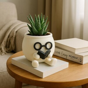 May include: White ceramic planter shaped like a person wearing glasses and holding a book. The planter has a green succulent plant growing inside. The planter is sitting on a stack of books with the text "Live Every Moment" on the top book.