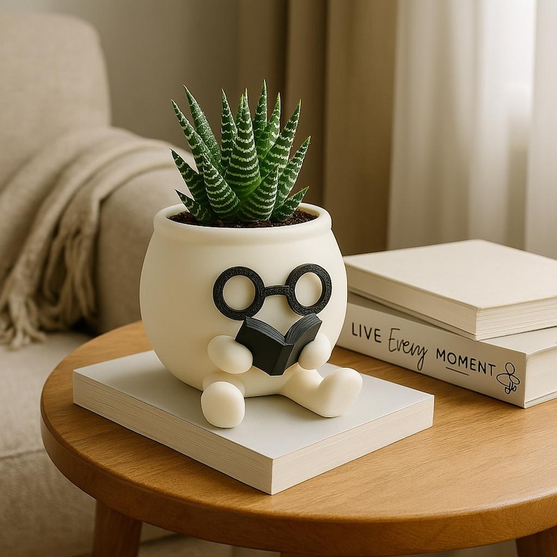 May include: White ceramic planter shaped like a person wearing glasses and holding a book. The planter has a green succulent plant growing inside. The planter is sitting on a stack of books with the text "Live Every Moment" on the top book.