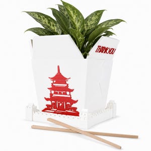 May include: A white takeout box planter featuring a red pagoda graphic on the front and the text THANK YOU on the side flap. The base is designed like a fortress wall, and the planter holds a green leafy plant with two wooden chopsticks in front.