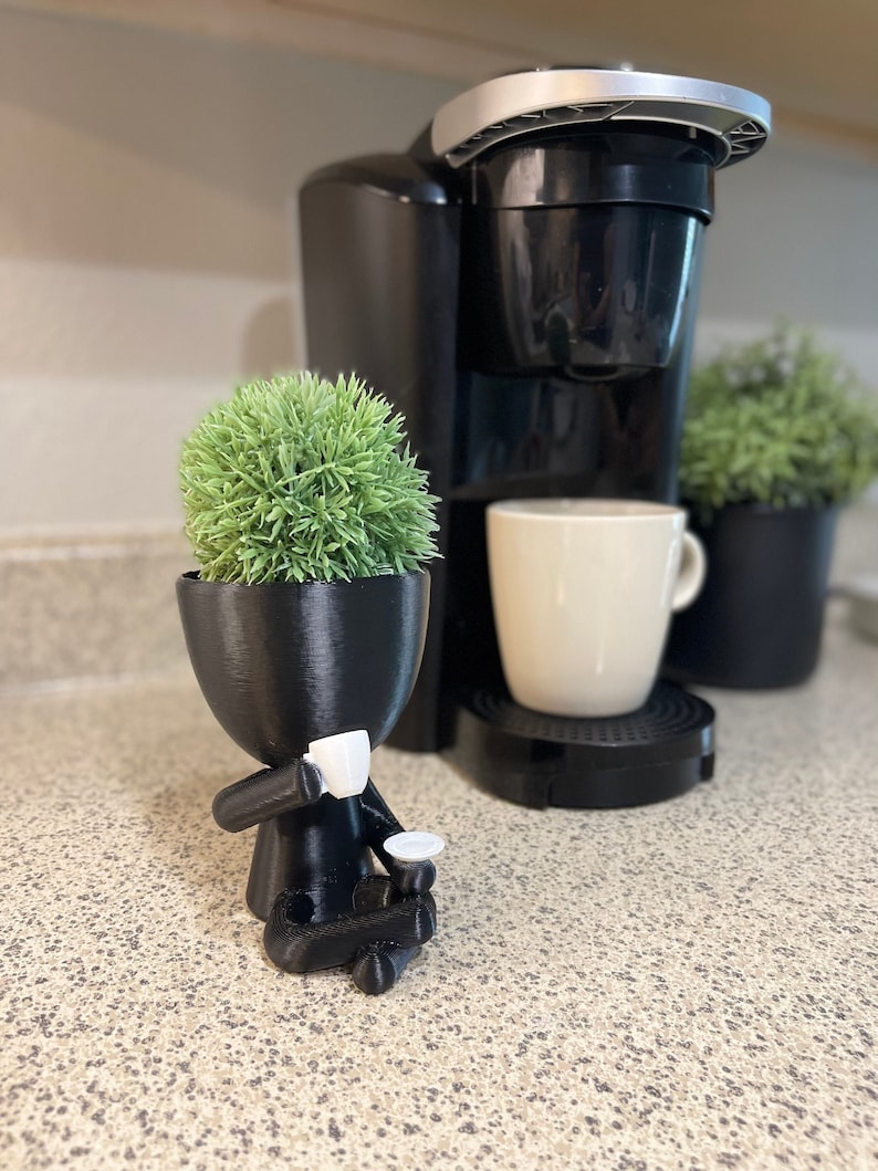 May include: A black 3D-printed planter shaped like a person, holding a small cup and saucer. The planter contains a green, spherical plant. A black coffee maker and a white mug are in the background, suggesting a coffee-themed decor.