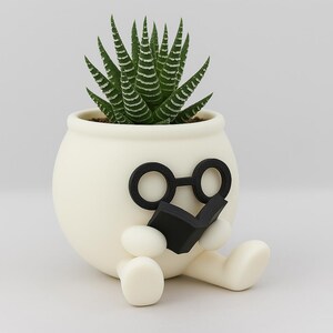 May include: White ceramic planter shaped like a person wearing glasses and reading a book. The planter has a green succulent plant growing inside.