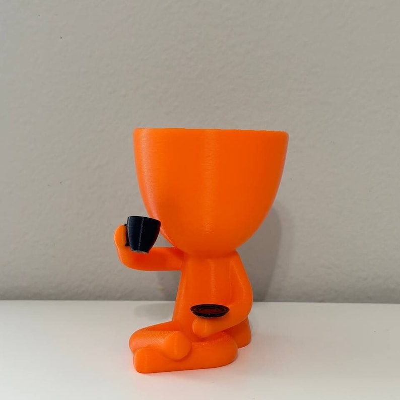 May include: An orange 3D-printed planter shaped like a person seated in a meditative pose, holding a black teacup and saucer. The planter has a large opening for plants. The figure is sitting on a white surface.