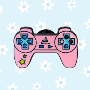 May include: A pink enamel pin shaped like a video game controller. The controller has a black outline, blue directional buttons, and colorful action buttons. A yellow star and a light blue background with white flowers complete the design.