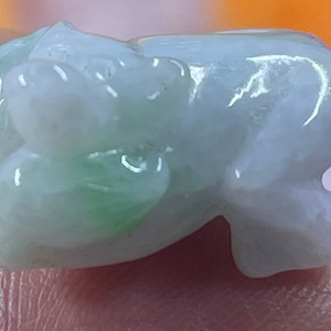 May include: A small, green and white jade carving of a pig. The pig is lying on its side with its head turned to the side. The carving is smooth and polished.