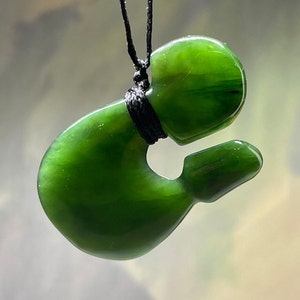 May include: A green jade pendant carved in the shape of a traditional Maori Hei Tiki, a symbol of good luck and protection. The pendant is strung on a black cord.