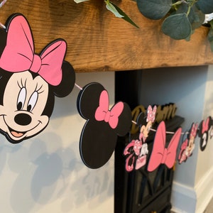 K&ouml;nnte beinhalten: Eine Minnie-Maus-Themenbanner mit schwarzen und rosa Ausschnitten. Das Banner zeigt Minnie Mouses Gesicht, Schleifen und andere charakterbezogene Designs. Das Banner ist mit rosa Schnur aufgereiht und eignet sich wahrscheinlich f&uuml;r eine Geburtstagsfeier.