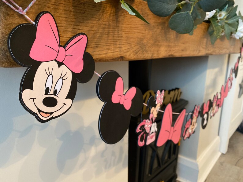 K&ouml;nnte beinhalten: Eine Minnie-Maus-Themenbanner mit schwarzen und rosa Ausschnitten. Das Banner zeigt Minnie Mouses Gesicht, Schleifen und andere charakterbezogene Designs. Das Banner ist mit rosa Schnur aufgereiht und eignet sich wahrscheinlich f&uuml;r eine Geburtstagsfeier.