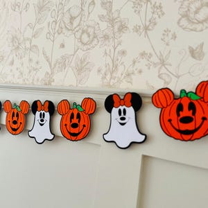 May include: A Halloween garland with Mickey and Minnie Mouse-themed pumpkin and ghost decorations. The pumpkins are orange with green stems, and the ghosts are white. The garland is set against a floral wallpaper background.