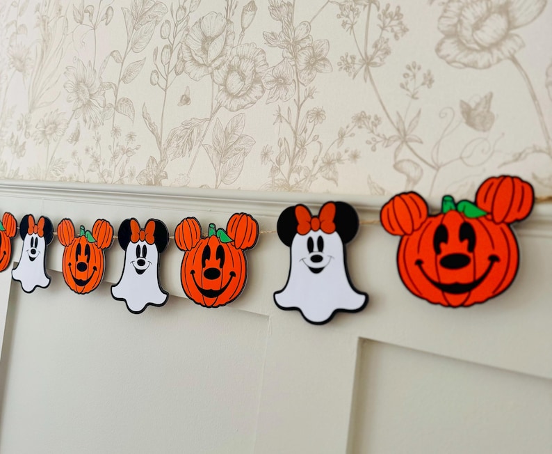 May include: A Halloween garland with Mickey and Minnie Mouse-themed pumpkin and ghost decorations. The pumpkins are orange with green stems, and the ghosts are white. The garland is set against a floral wallpaper background.