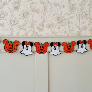 May include: A Halloween garland with orange pumpkin and ghost-shaped decorations with black detailing. The garland is strung across a white wall, creating a festive display for the holiday. The garland is a decorative item.