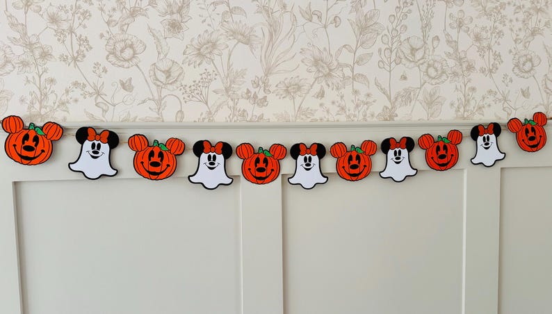 May include: A Halloween garland with orange pumpkin and ghost-shaped decorations with black detailing. The garland is strung across a white wall, creating a festive display for the holiday. The garland is a decorative item.