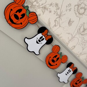May include: A Halloween garland with orange pumpkin and ghost-shaped decorations. The pumpkin decorations have smiling faces and green stems. The ghost decorations have Minnie Mouse features, including a red bow. The garland is strung on natural twine.