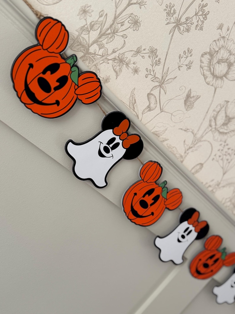 May include: A Halloween garland with orange pumpkin and ghost-shaped decorations. The pumpkin decorations have smiling faces and green stems. The ghost decorations have Minnie Mouse features, including a red bow. The garland is strung on natural twine.