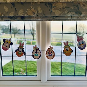 May include: An Easter-themed banner with Disney characters, such as Mickey and Minnie Mouse, Chip and Dale, Donald Duck, and Daisy Duck, each wearing bunny ears. The garland is strung across a window overlooking a green field.