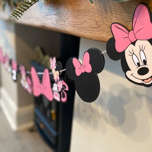 K&ouml;nnte beinhalten: Eine dekorative Girlande mit Minnie Mouse. Die Girlande enth&auml;lt Ausschnitte von Minnie Mouses Kopf, Schleifen und andere Charakterelemente. Das Farbschema ist Schwarz, Rosa und Wei&szlig;, zusammengef&uuml;gt mit wei&szlig;er Schnur. Ideal f&uuml;r eine Party oder Feier.
