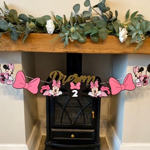 K&ouml;nnte beinhalten: Eine Minnie-Mouse-Geburtstagsgirlande mit rosa Schleifen, Minnie-Mouse-Ausschnitten und der Zahl 2. Die Girlande ist &uuml;ber einem h&ouml;lzernen Kaminsims mit Gr&uuml;n und wei&szlig;en Blumen gespannt. Das Wort "Dream" ist ebenfalls zu sehen.