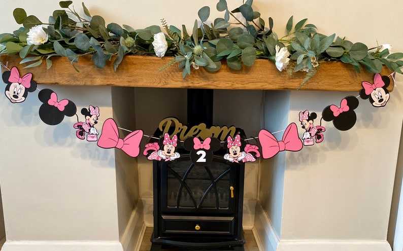 K&ouml;nnte beinhalten: Eine Minnie-Mouse-Geburtstagsgirlande mit rosa Schleifen, Minnie-Mouse-Ausschnitten und der Zahl 2. Die Girlande ist &uuml;ber einem h&ouml;lzernen Kaminsims mit Gr&uuml;n und wei&szlig;en Blumen gespannt. Das Wort "Dream" ist ebenfalls zu sehen.