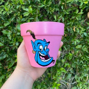 Könnte beinhalten: Ein rosa Terrakotta-Blumentopf mit einer handgemalten Illustration des Dschinni aus Disneys Aladdin. Der Dschinni ist blau mit einem verschmitzten Grinsen und einem goldenen Ohrring.