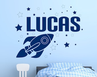 Pode incluir: Adesivo de parede azul com foguete e estrelas com o nome "LUCAS" em letras em negrito. O adesivo está em uma parede azul claro com uma cama e outros móveis no fundo.