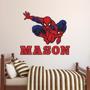 Puede incluir: Un adhesivo de pared Spiderman rojo y azul con un dise&ntilde;o de telara&ntilde;a. El adhesivo est&aacute; sobre una cama de madera marr&oacute;n con una manta a rayas. El nombre "MASON" est&aacute; escrito en letras rojas con un dise&ntilde;o de telara&ntilde;a.