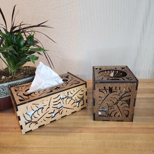 Puede incluir: Dos cubiertas de caja de pañuelos de madera con un diseño de hojas tropicales. Las cubiertas son de madera oscura y tienen un diseño de hojas recortadas. Las cubiertas están sobre una superficie de madera.