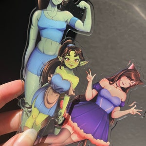 May include: Three clear acrylic charms featuring fantasy characters. The characters are depicted in various poses and outfits, including a blue crop top and shorts, a blue dress with a pink trim, and a blue dress with a brown apron.