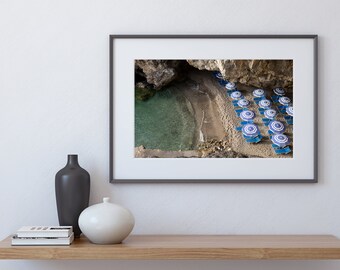 May include: An aerial view of a secluded beach with turquoise water and a rocky cliff.  The beach is lined with blue and white striped beach umbrellas and lounge chairs.
