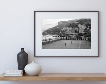 May include: Black and white photograph of a group of children playing soccer on a court with a net. The court is located near the ocean and a hillside with buildings.
