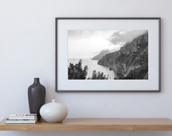 May include: A black and white photograph of a rocky coastline with a body of water in the distance. The photo is framed in a gray frame and is hanging on a white wall.