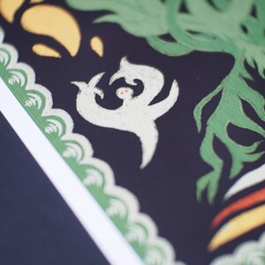 May include: Close-up of a decorative art print with a black background. The print features white, green, yellow, and orange stylized shapes. A white figure with red dots is in the center, surrounded by green foliage.
