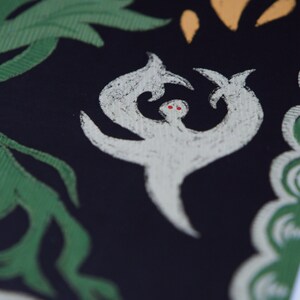 May include: Close-up of a black textile with a white, stylized figure with red eyes. Green leaves and yellow shapes are in the background. The figure has a unique, abstract design.