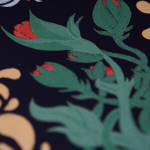 May include: Close-up of a floral design with green leaves and red flower buds against a black background. The design also includes white, yellow, and red decorative elements. The image has a textured, hand-painted appearance.